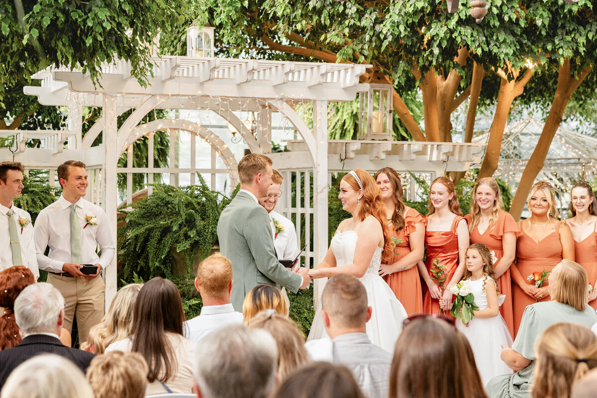 The bride and groom exchange vows under a white pergola adorned with fairy lights, surrounded by their bridal party and guests at Le Jardin in Sandy, Utah.