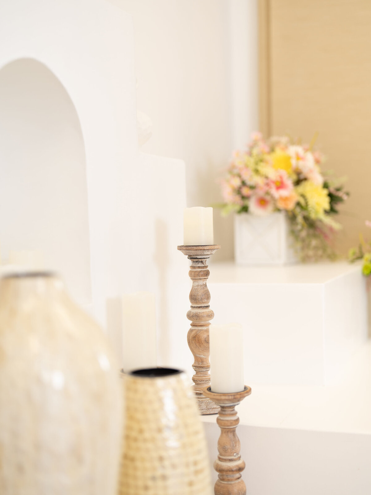 Wooden candlesticks with unlit white candles stand on a white surface, accompanied by soft wicker vases. A blurred pastel flower bouquet in the background adds a serene touch.