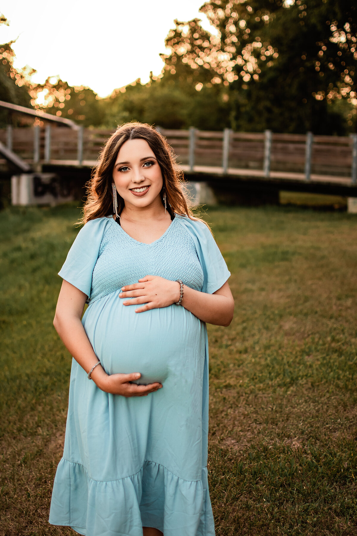 A mom to be wearing a blue dress holds her bump and smiles art the camera.