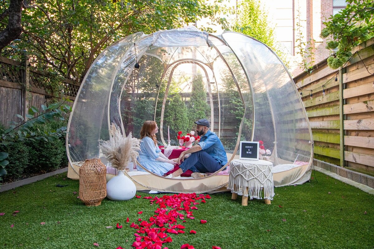 A romantic backyard picnic setup inside a clear dome tent, styled with rose petals, candles, and boho décor by Yours Truly, a New Jersey wedding planner and designer.