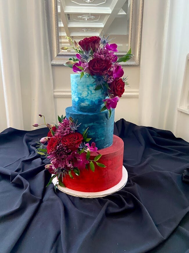The photo shows a three tier cake sitting on a table with a black tablecloth. The cake has marbled textured frosting in (from top to bottom) teal, blue, and reds. Its also decorated with fresh flowers in pink, purple, and red jewel tones. 