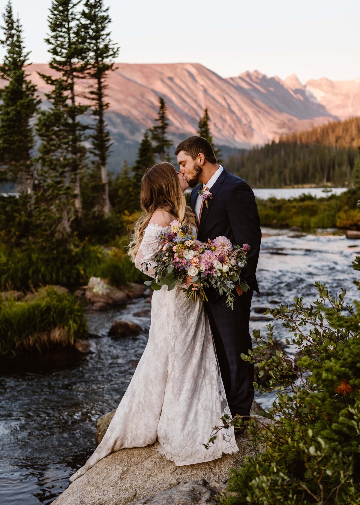 Couple kisses on a rock with alpenglow lighting up the mountains behind them at sunrise during their Lake Isabelle elopement