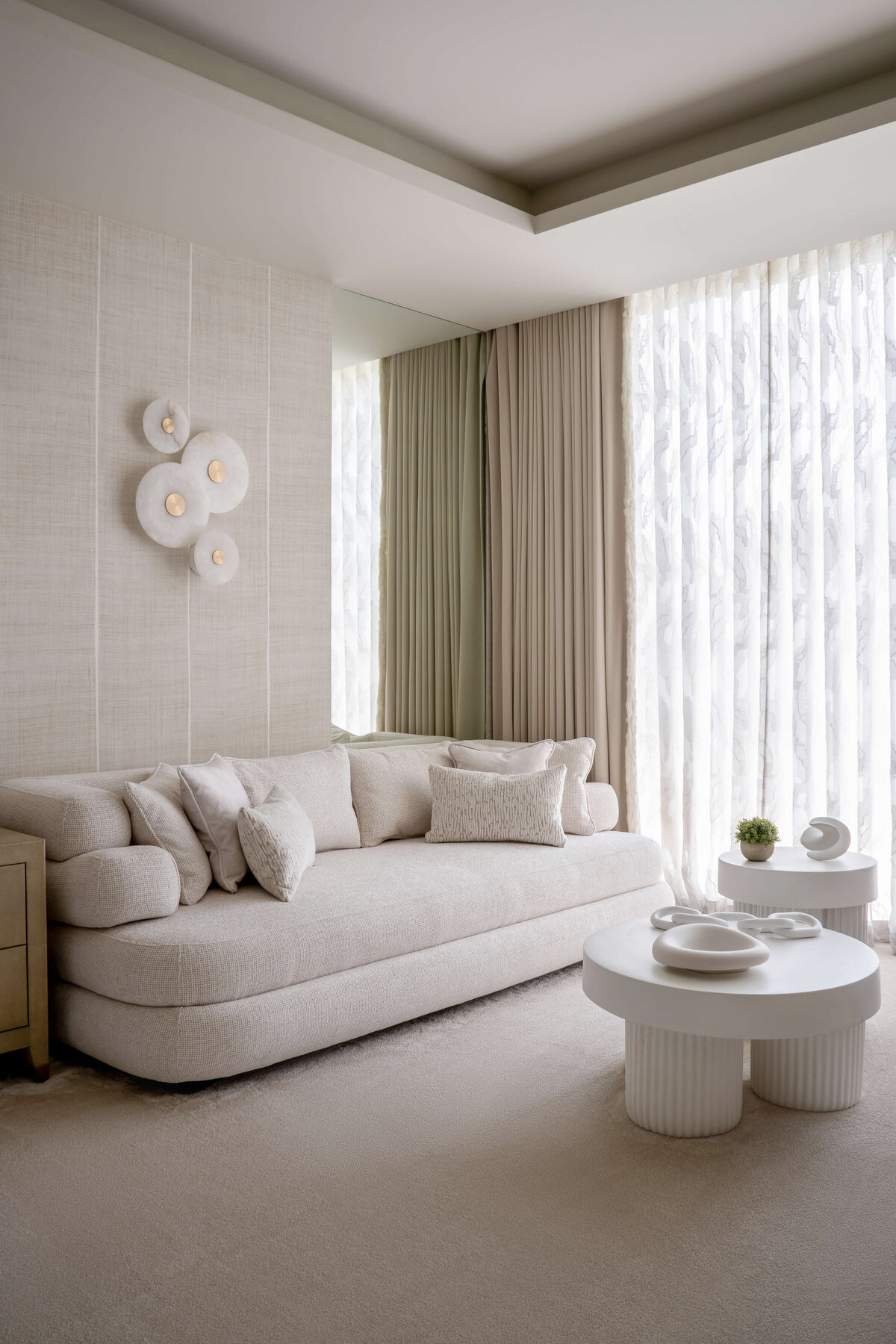 A sitting area in a airy, neutral primary bedroom featuring a large white couch and a contemporary white coffee table.