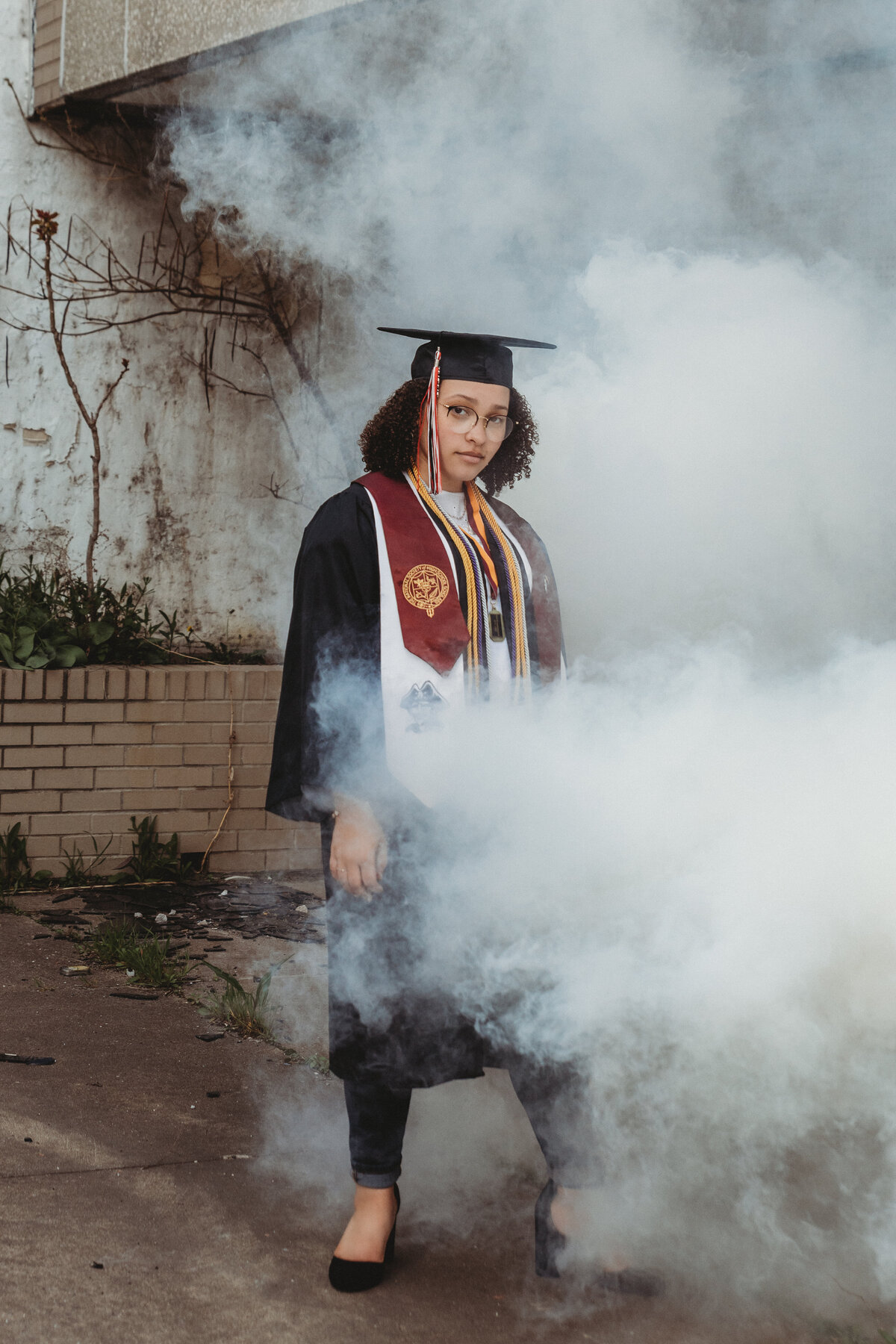 Graduate dressed in gown stands amongst smoke at abandoned location in Cleveland.