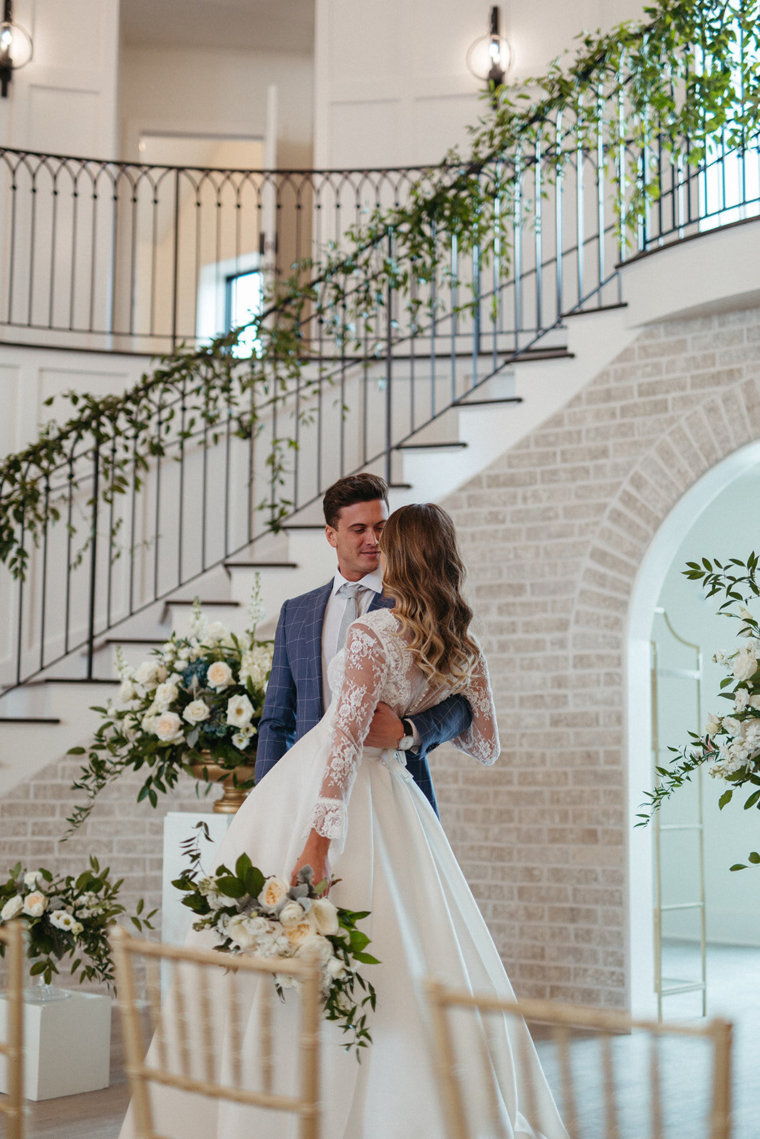 A groom in a blue suit holds bride wearing a white wedding gown and holding a bouquet in front a staircase wrapped in garland.