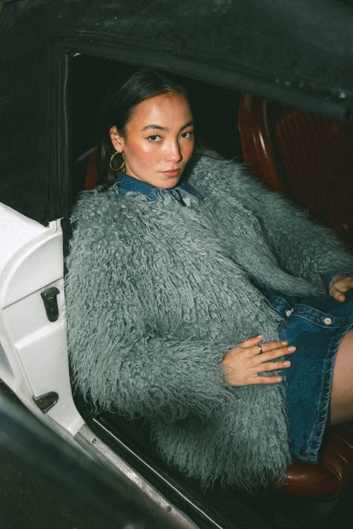 A close-up of the model resting inside the vintage car, her expression calm and thoughtful, wrapped in a textured fur coat. The shot captures an intimate moment, highlighting the softness of the coat against the sleek interior of the car. Photographed by Bo Eppink.