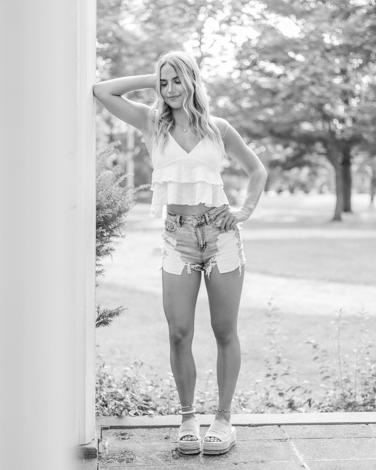 Black and white photo of a senior high school female, wearing blue jean shorts, spaghetti strap shirt, standing on a porch, hand on her hip, looking down with a soft smile.