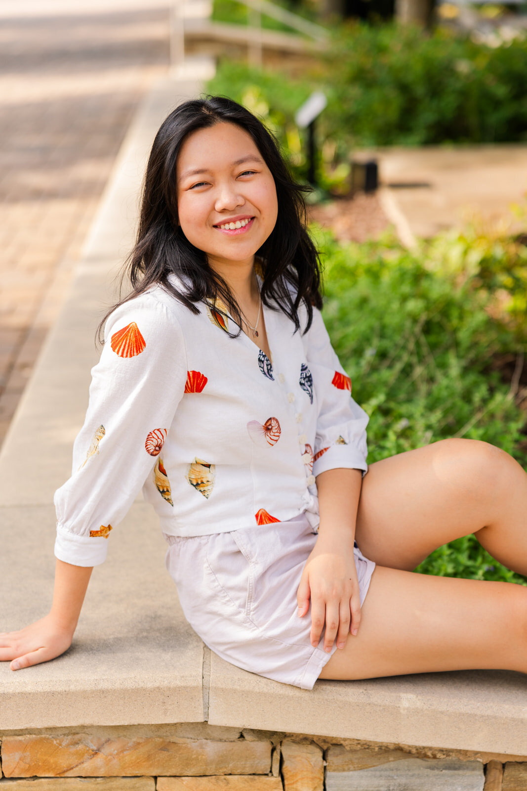 senior girl sitting legs stretched turned sideway towards the camera with a mix or green and city background during photo session