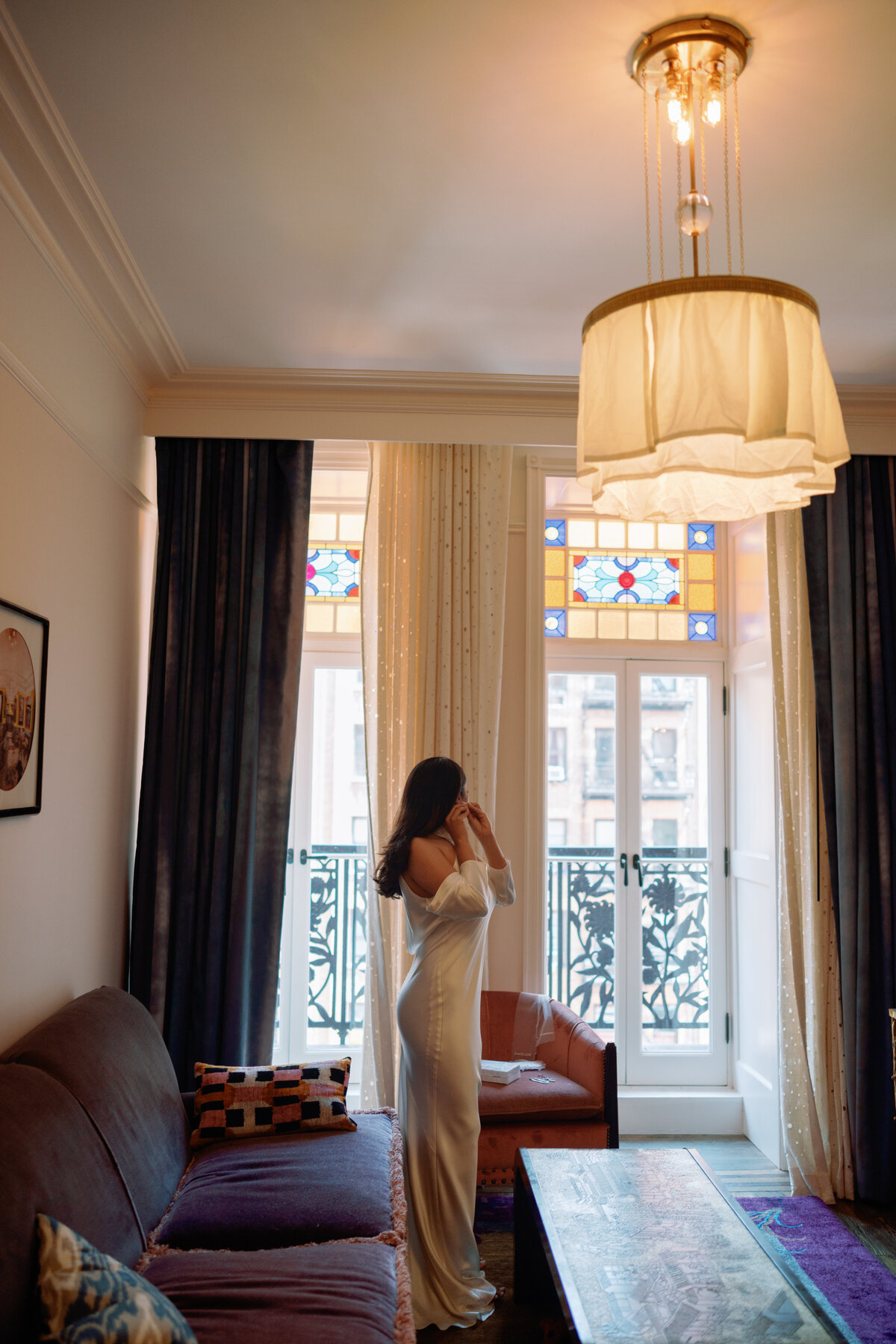 Bride in an elegant white gown standing by the window with stained glass light at Hotel Chelsea in New York City, captured during Japna and Chris’s intimate elopement by NYC wedding photographer Perry Hancock.