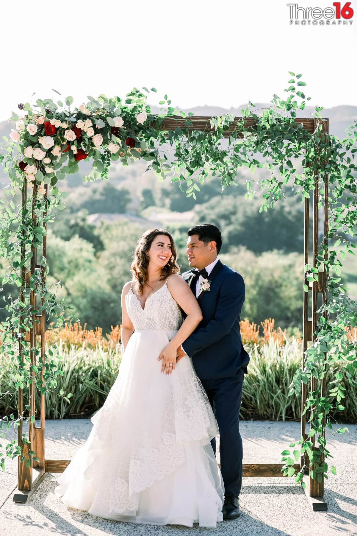 Bride looks back at her Groom as he holds her waist while posing under the floral archway