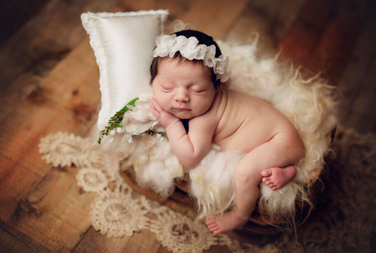 A newborn baby sleeps peacefully on a fluffy, woolen blanket during an in-home newborn session in Central Florida. The baby wears a floral headband and rests beside a small white pillow, with delicate lace decorating the area, capturing the essence of Orlando newborn photography.