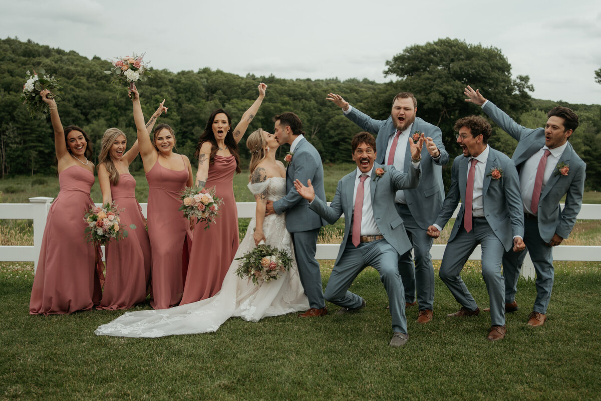 A newlywed couple shares a kiss while surrounded by their excited bridal party. The bride wears a beautiful white gown with a long train, and the bridesmaids are in matching dusty rose dresses holding bouquets of pink and white flowers. The groom and groomsmen are dressed in light blue suits with pink ties, showing their excitement with joyful expressions and animated poses. The group stands on a lush green lawn with a scenic backdrop of trees, creating a lively and celebratory wedding day moment.