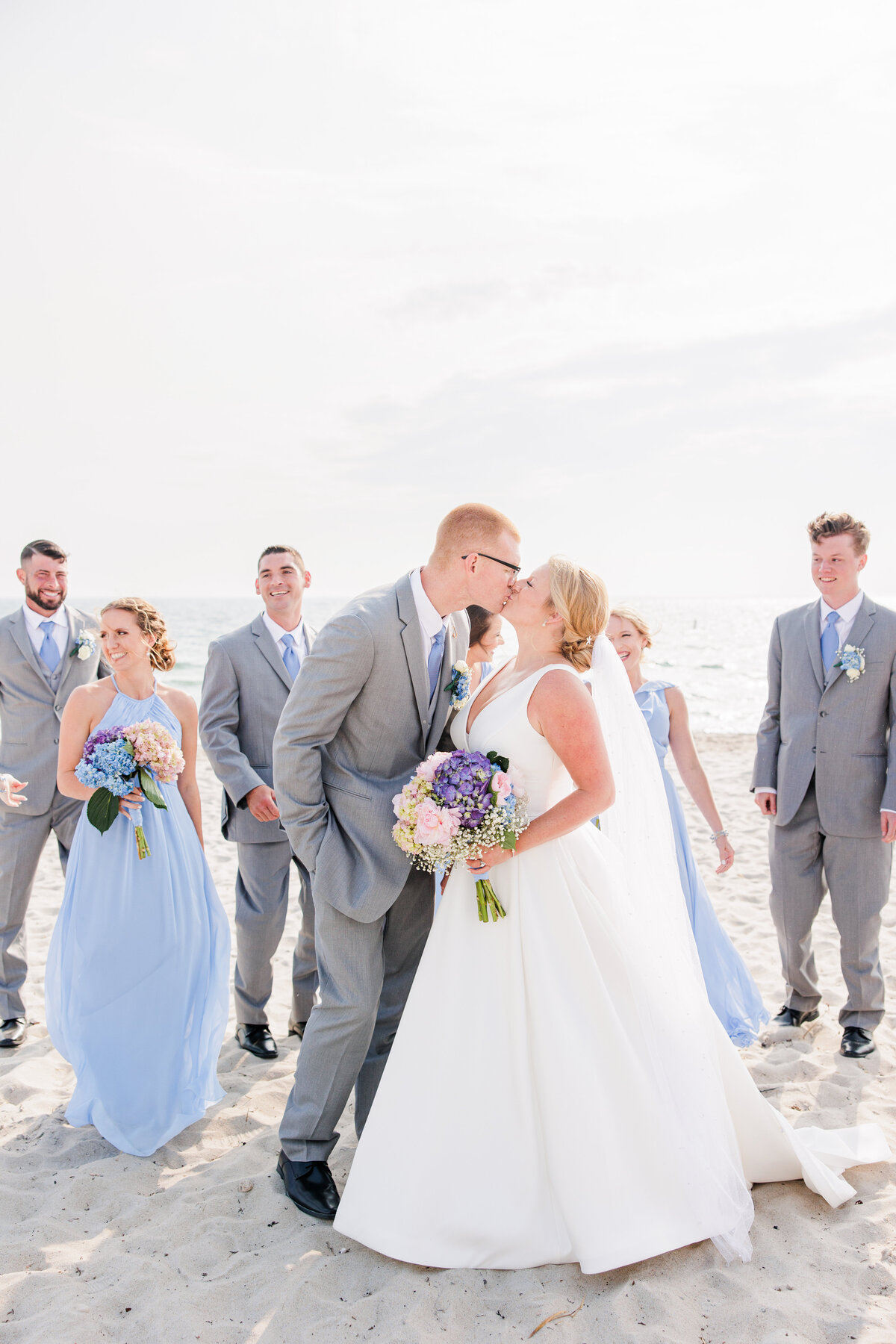 A bride and groom kissing while their wedding party laughs representing a joyful Cape Cod wedding photographer