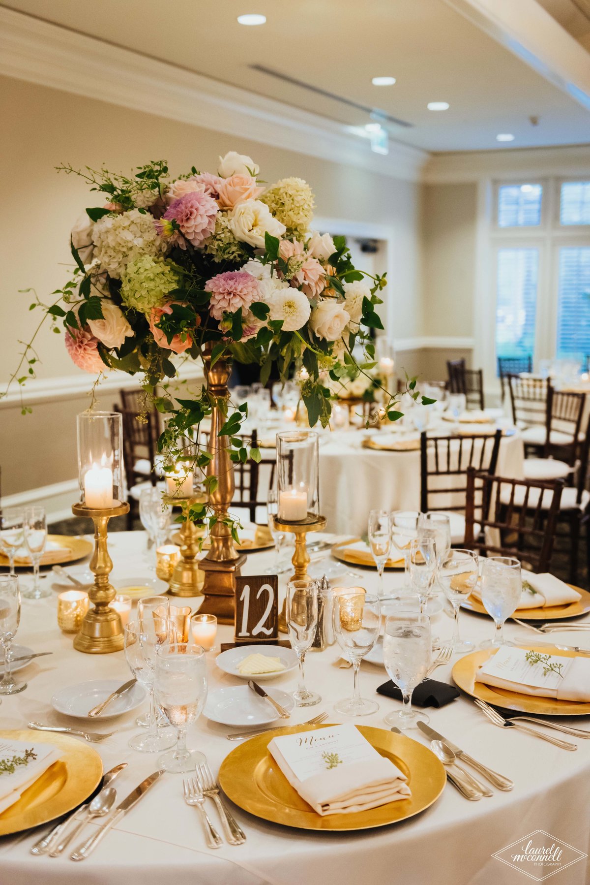 elevated centerpiece of peach dahlias, peach and ivory roses, trailing greenery on gold stand on table with ivory linen and gold charger plates