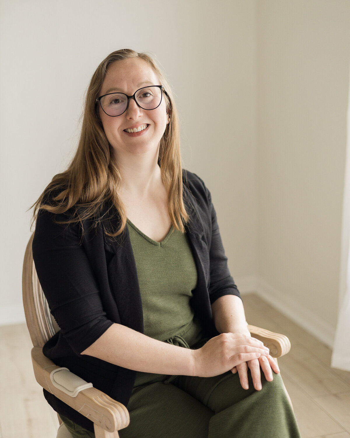 Strawberry blonde female, sitting in a chair, wearing a dark green shirt and pants with a black jacket over the green outfit, wearing glasses, smiling at the camera