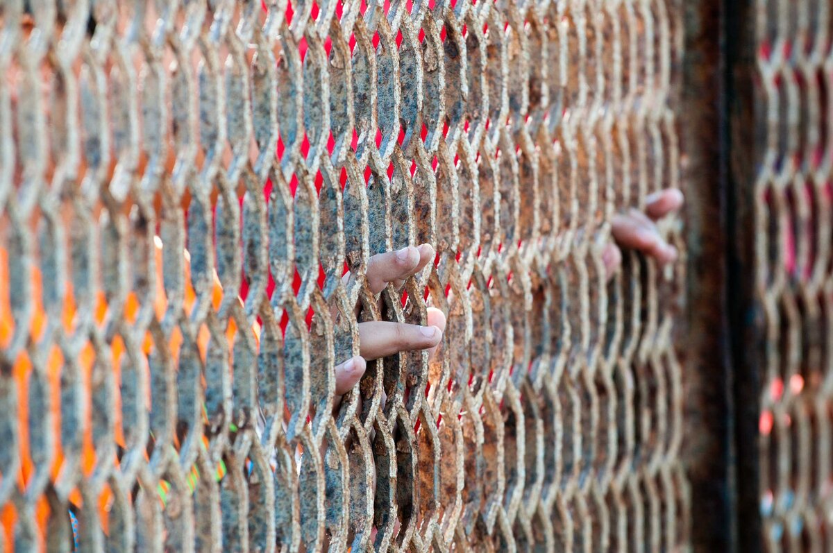 Fingers are shown poking through the rusted border fence at friendship park