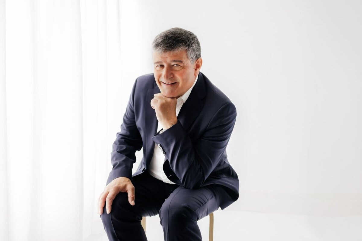 Man in blue suit seated on stool, chin on fist, smiling for professional portrait by Ruaa Hamdan Photography, Altrincham