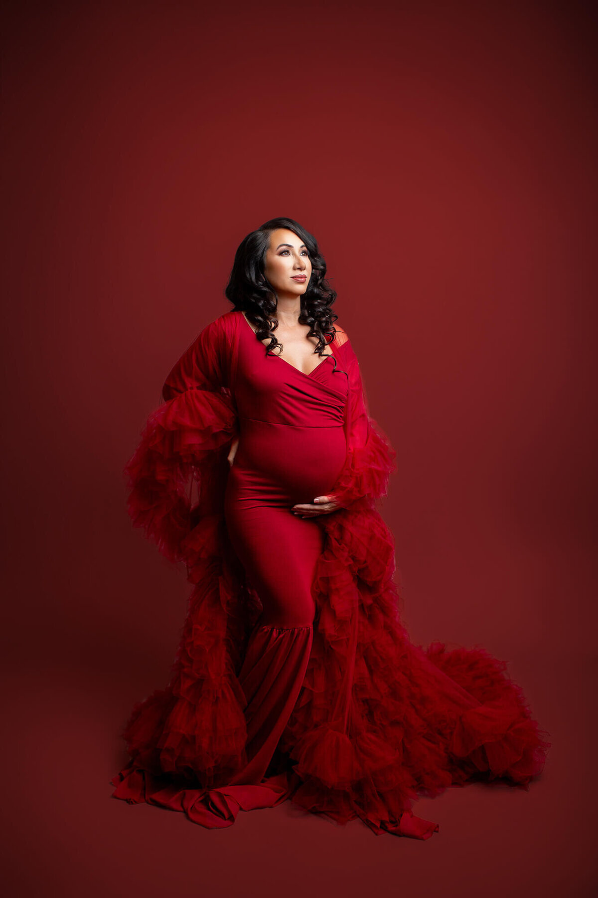 Maternity session in studio with mother in dramatic red dress looking up to light on red background 