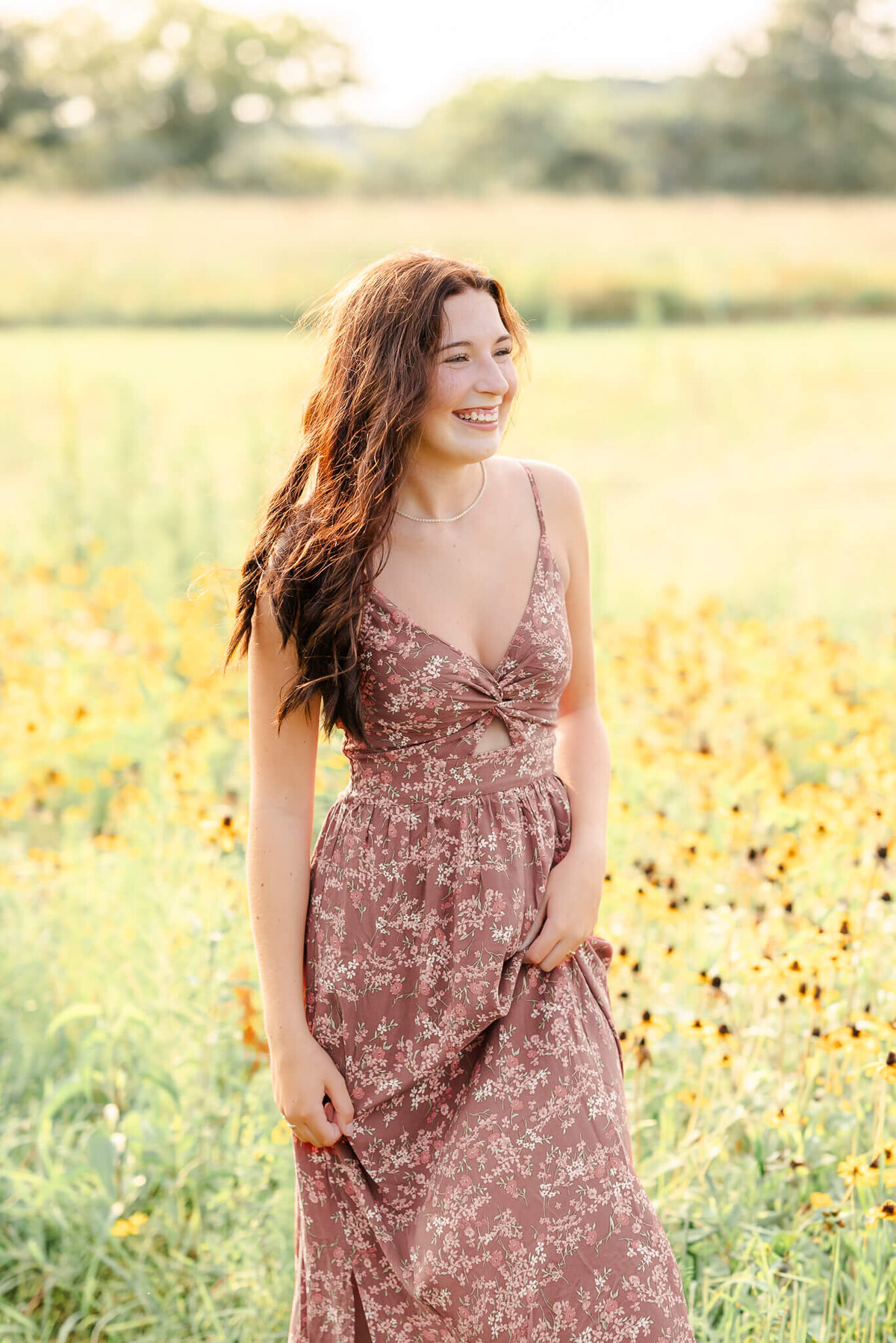 A high school senior, wearing a long floral dress, walks among some yellow flowers while laughing.