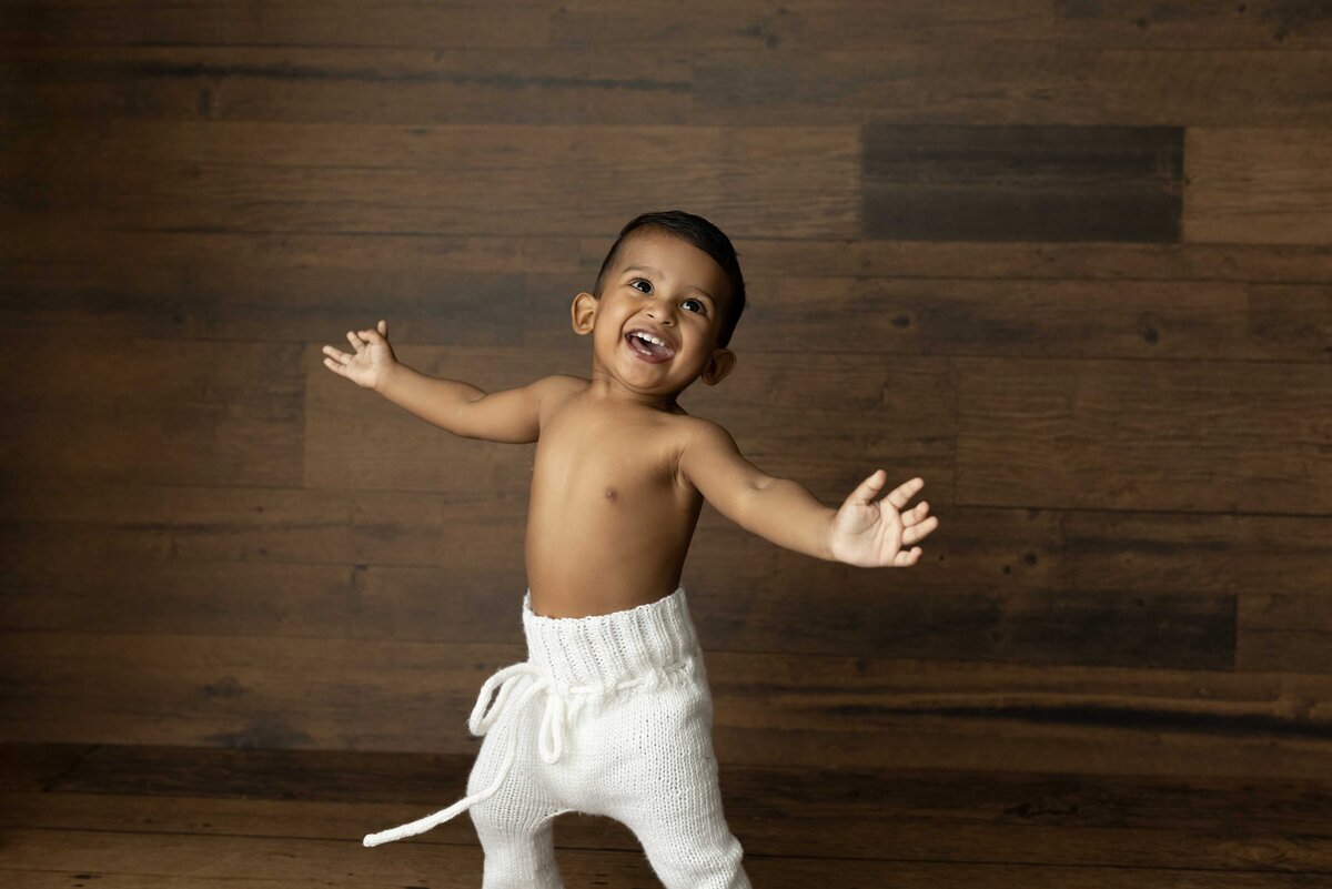 Happy baby in white knit pants dancing during London, Ontario photo session. In the background is a wood wall.