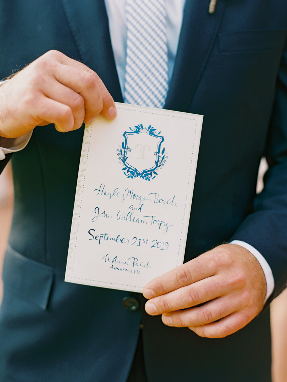 The photo was captured at The Hamptons, New York, during a waterfront estate tented wedding by acclaimed photographer Bonnie Sen. Additionally, East West Films videographers were present to capture the beautiful moments of the event. In the image, a person in a dark suit is seen holding a wedding program showcasing a beautiful blue crest with the initial 'T' and adorned with elegant calligraphy text. The program reveals the names Huxley Morgan Bosch and John William Torpey, along with the date of their nuptials, September 21st, 2019, at St. Anne's Parish in Annapolis, MD.