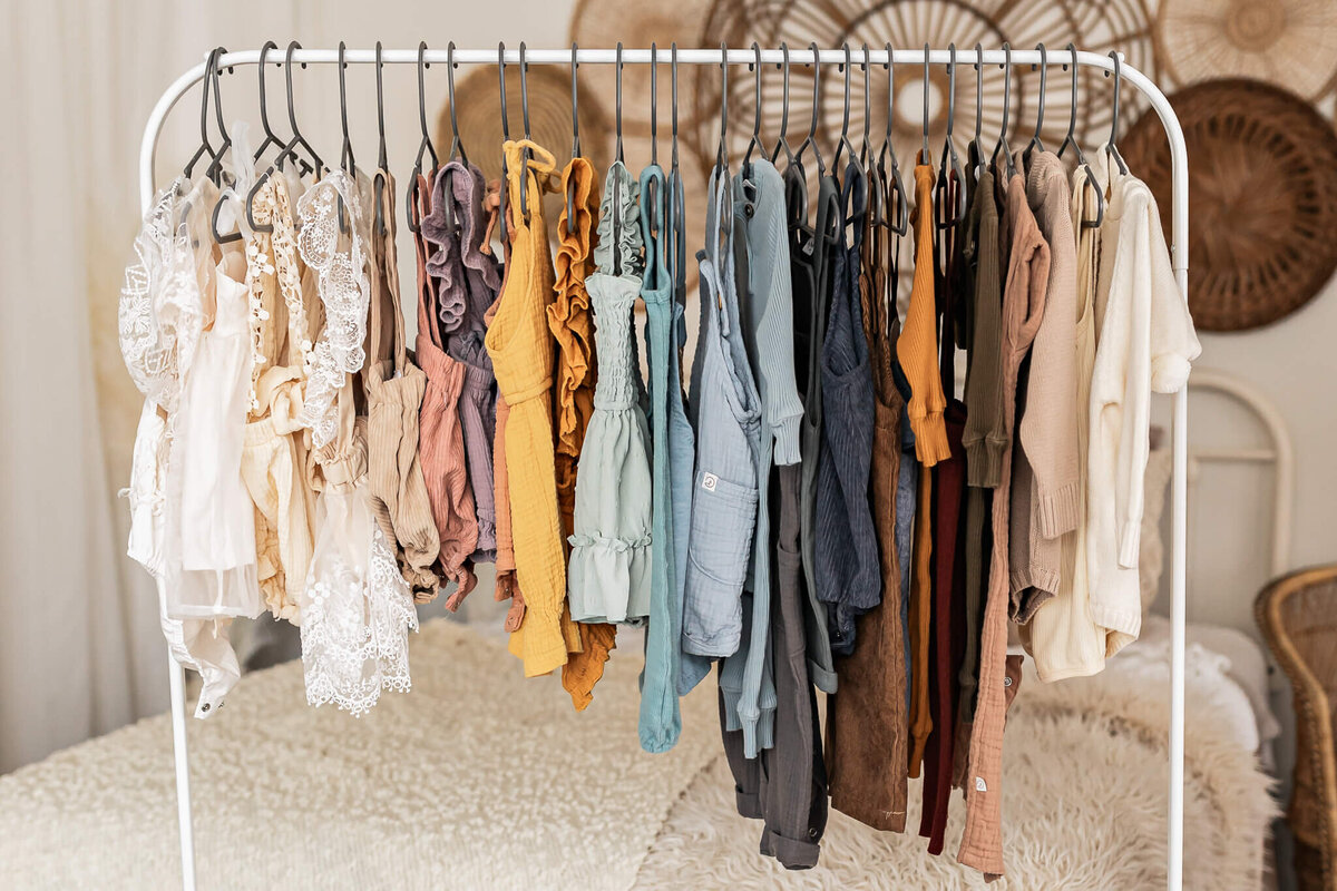 A clothing rack displays various colorful garments, including dresses and tops in shades of beige, pink, yellow, green, blue, and brown. The clothing features a mix of textures and styles, all arranged neatly on black hangers.