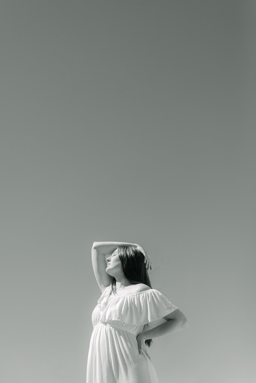 An airy black-and-white portrait of a woman in a flowing white dress, standing with her arm overhead against a clear sky, captured in a refined editorial style by Elizabeth Kane Photography—serving Philadelphia, New York, and DC.