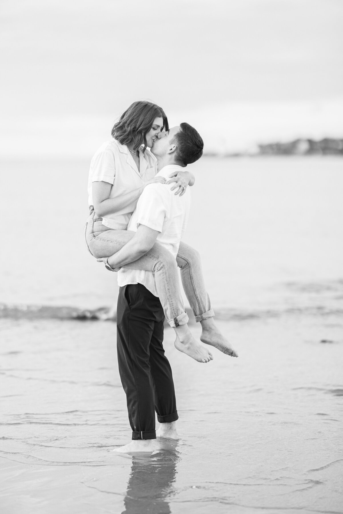 A black and white image of a man picking up a woman and sharing a kiss as they stand in the waves at Wingaersheek Beach during their sunset engagement photos.