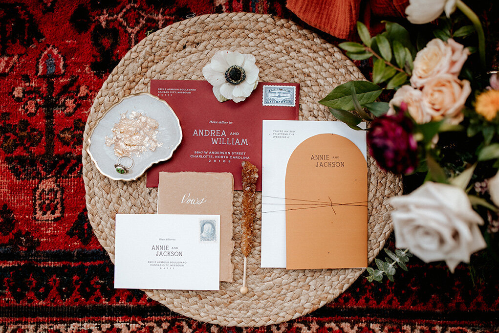 White and red wedding envelopes on circular woven basket on a red rug next to bouquet of flowers