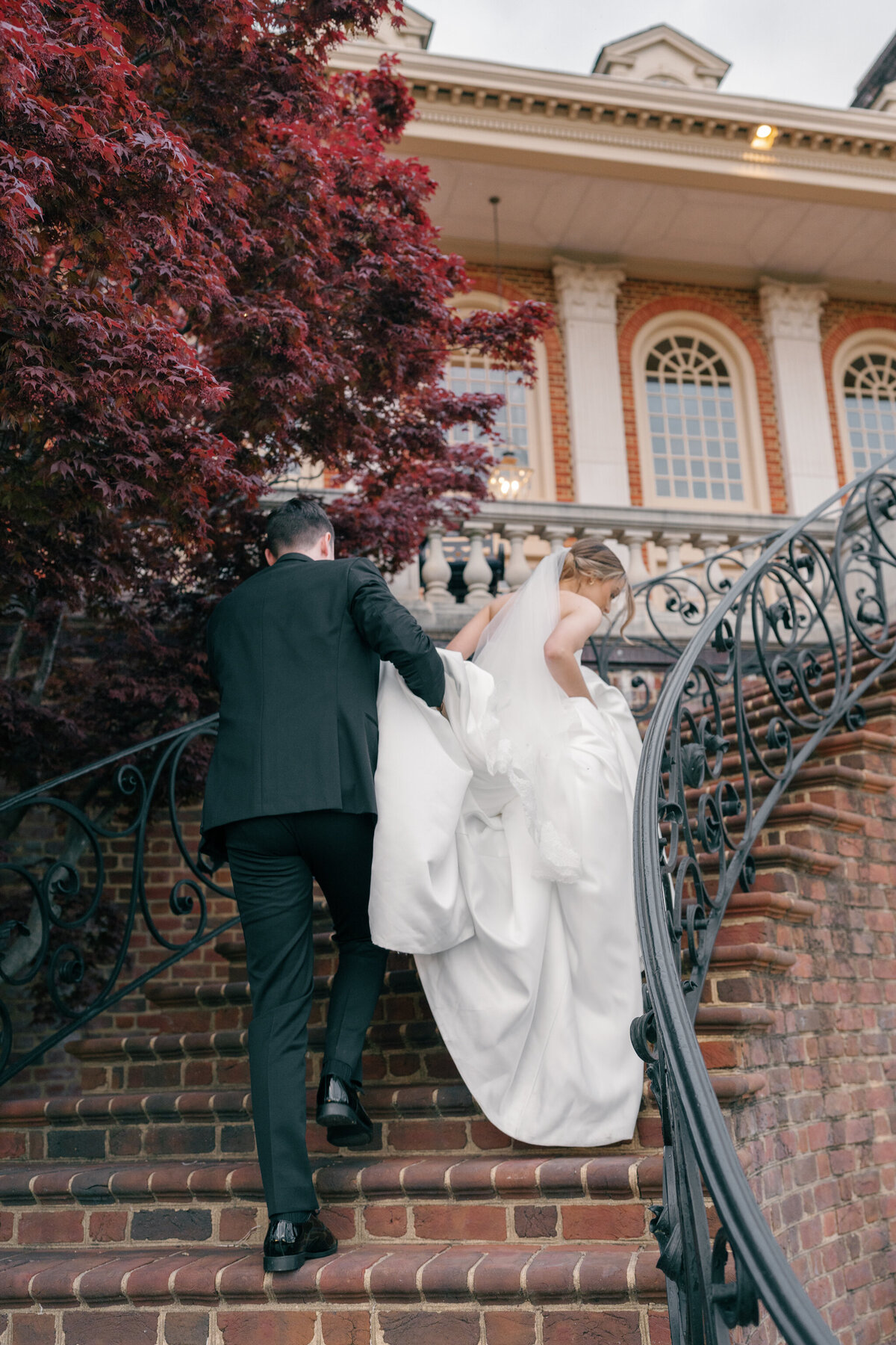 A groom tenderly lifts his bride’s gown as they ascend the grand brick staircase at The Estate at River Run. The intricate wrought iron railing and stately Georgian architecture frame this elegant moment, while the deep red foliage adds a touch of romance. Captured by Caitlin Wilcox Photography, a Richmond, VA film wedding photographer, this image embodies timeless sophistication and devotion.