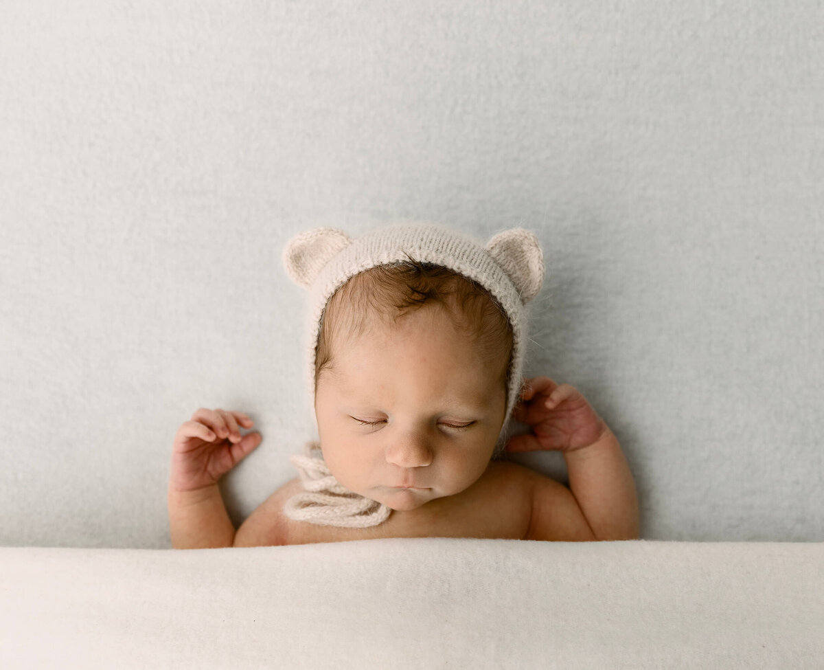 Photo of a newborn baby boy laying on a white backdrop with a teddy bear bonnet in an Erie photography studio