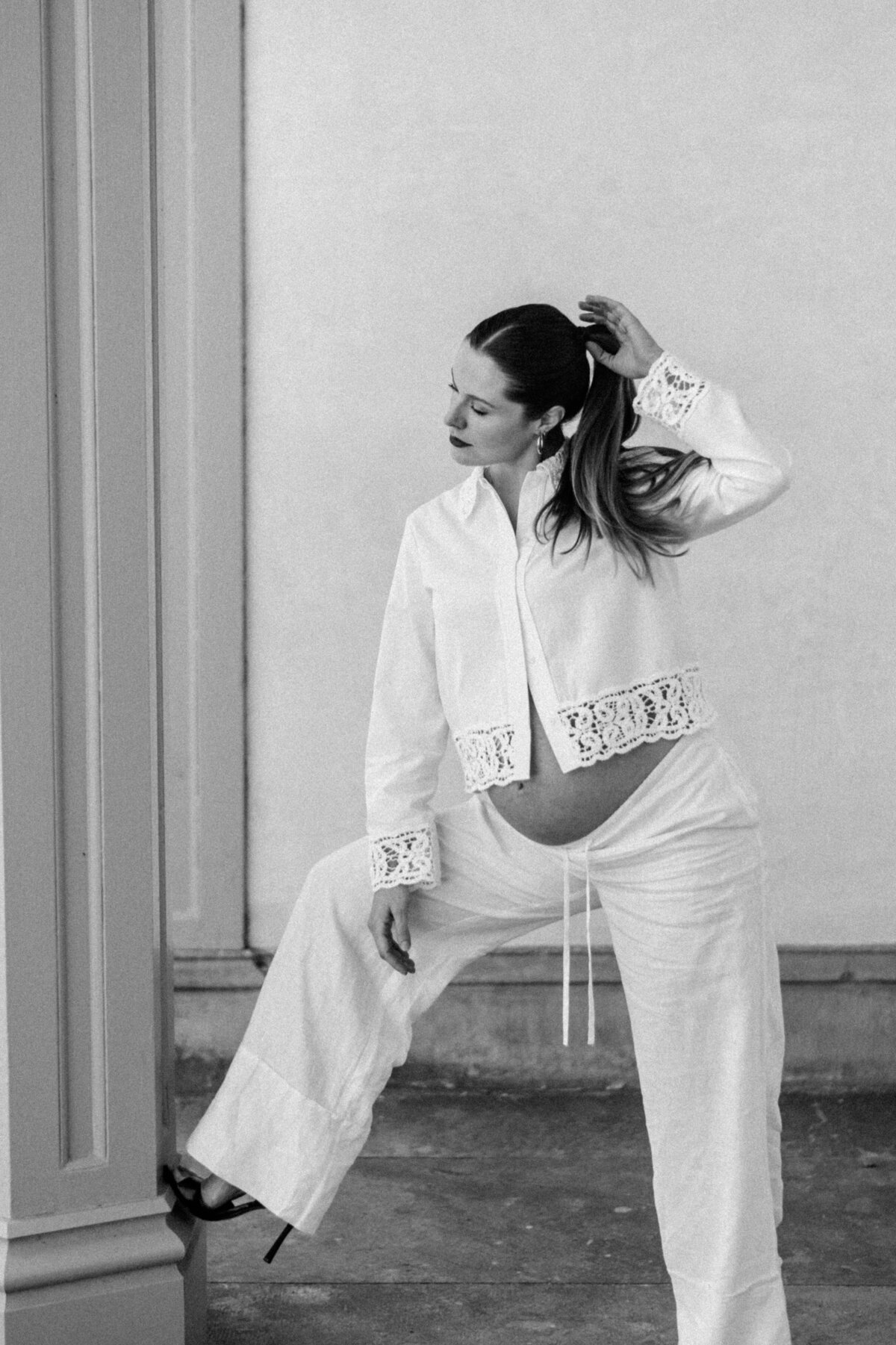 Editorial black-and-white maternity portrait of a poised mother-to-be in heels, framed by architectural columns