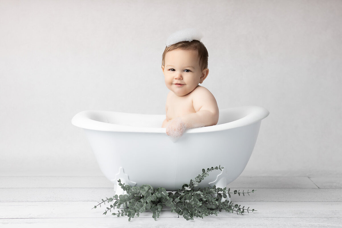 Milestone photo by DFW baby photographer showing a little one in a white clawfoot tub, wearing a crown of bubbles and smiling sweetly against a soft neutral backdrop.