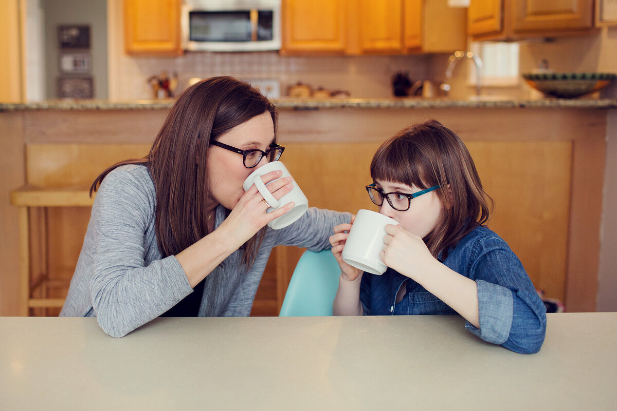 Mom and daughter look at each other as they both take a drink of hot chocolate.