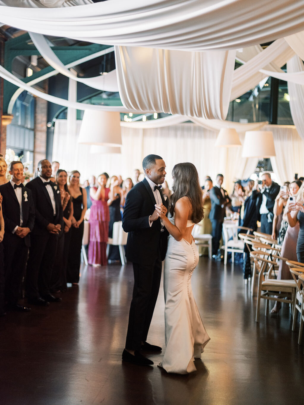 The bride and groom share their first dance surrounded by guests in an elegant, candlelit reception hall.