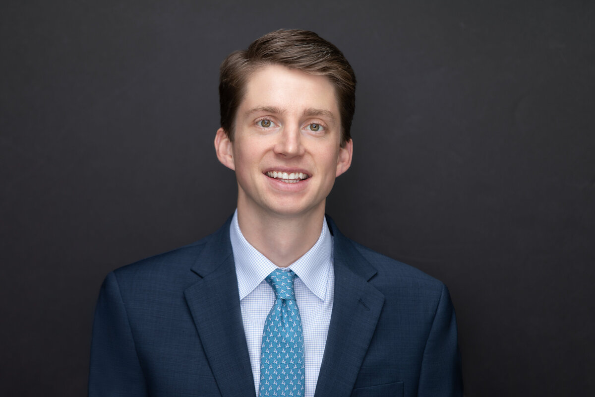 A smiling young businessman in a blue suit and light blue tie poses for a professional headshot on a black backdrop for Janel Lee Photography studios in Cincinnati Ohio