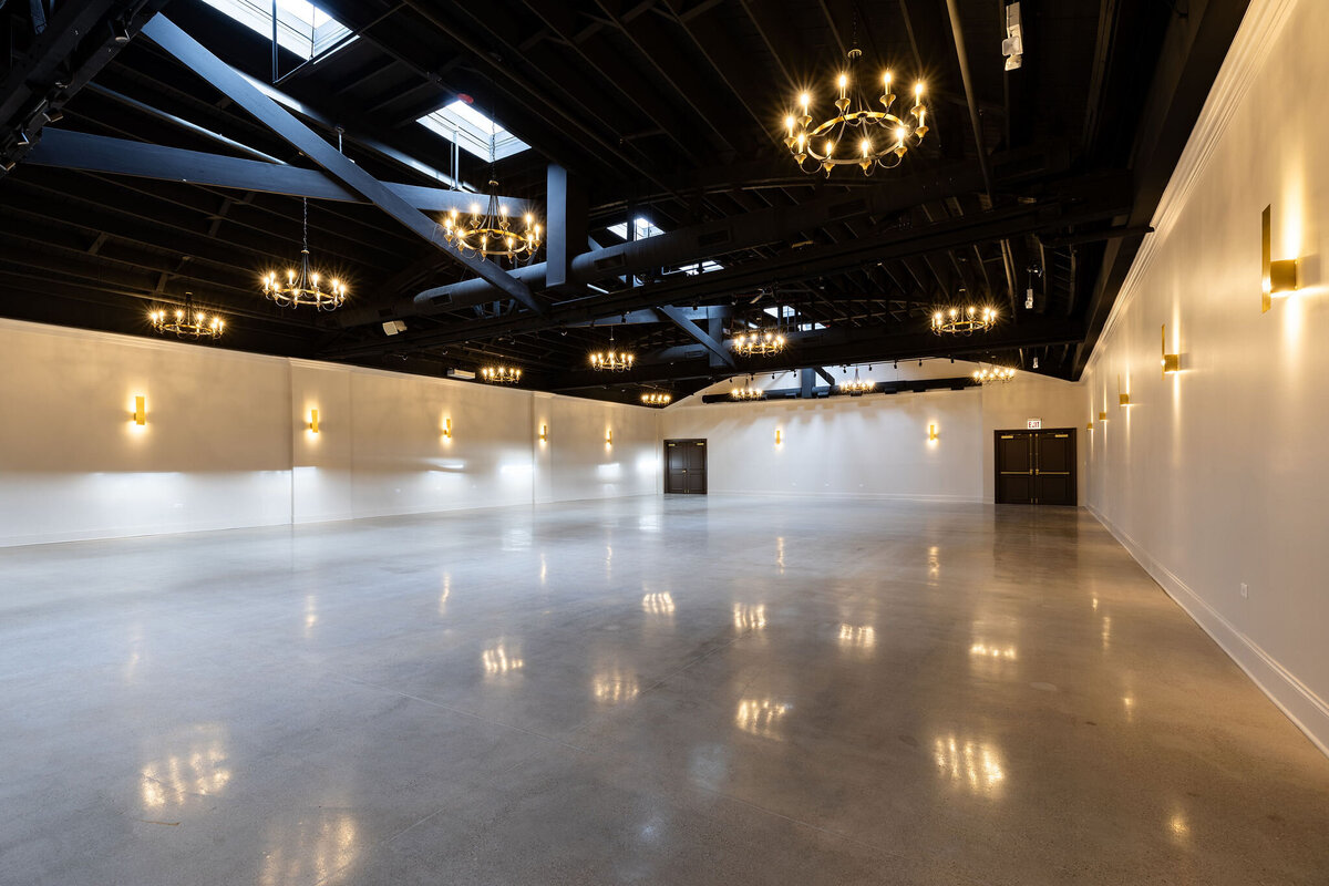 Spacious ceremony, reception or event space, with high industrial ceilings, polished floors, neutral walls and details, and ample natural light streaming through skylights and all around sconces and chandeliers, ready to be set up for an event