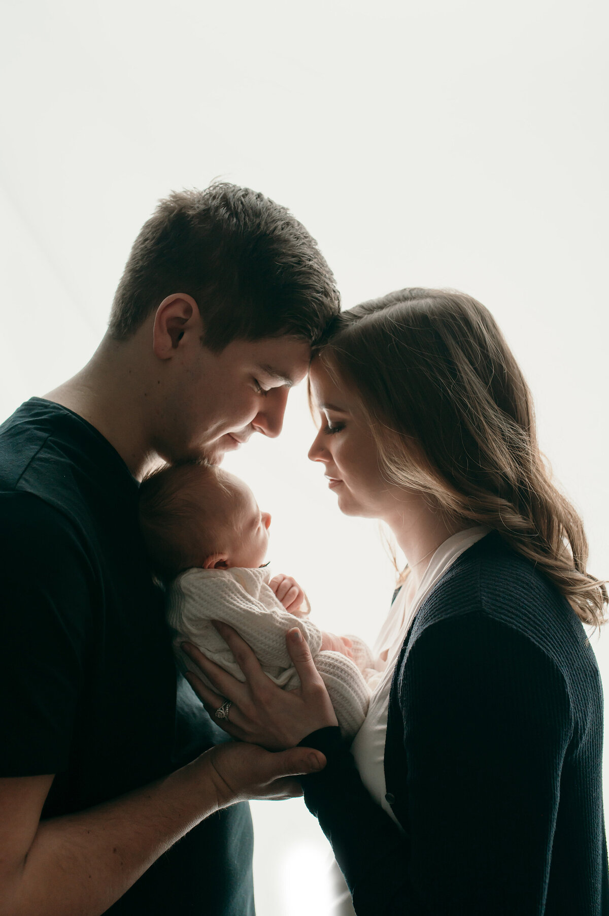 Portrait of Mom, Dad and infant shown in shadowed profile in front of a bright, white background. The parents are facing eachother with the baby held between them facing mom.