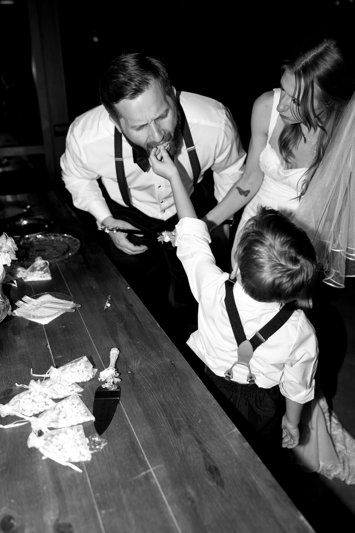 A groom eating caking from his sun while him and his new wife cut the wedding cake. 