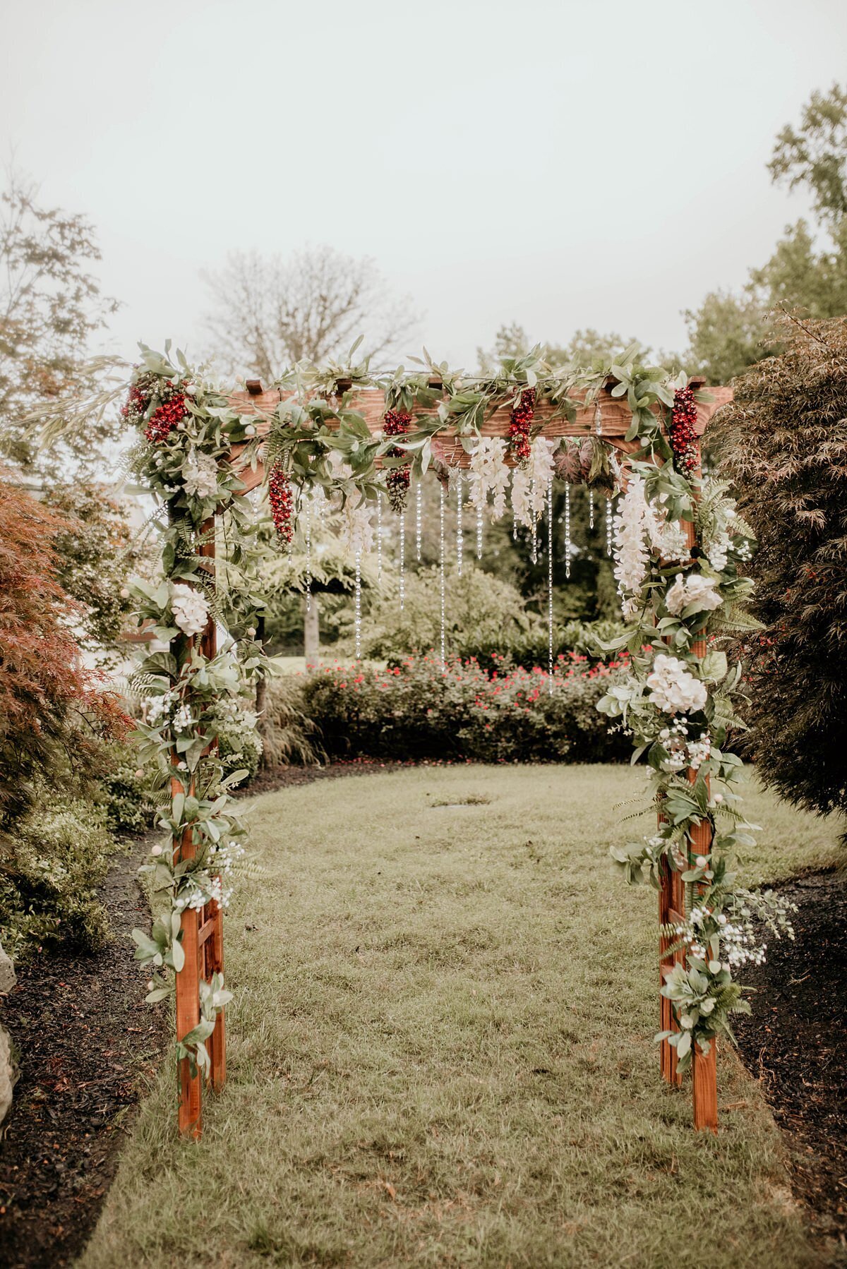 Wooden arbor set in the garden at The Estate at Cherokee Dock wrapped with greenery, hanging white flowers and hanging burgundy flowers accented by a curtain of crystals.