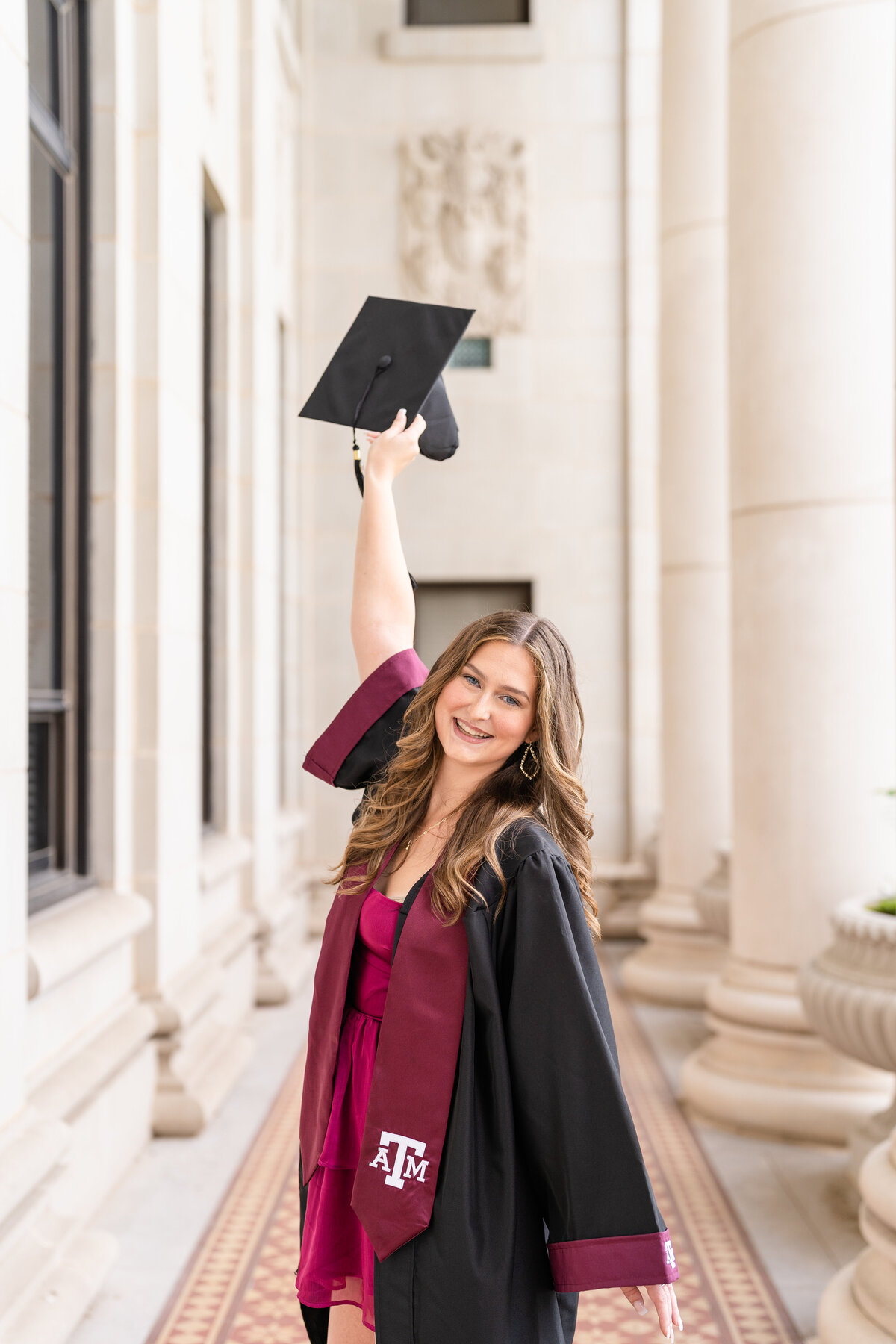 Texas A&M senior girl holding up grad cap while wearing gown and maroon stole and maroon dress and celebrating in middle of columns of Administration Building