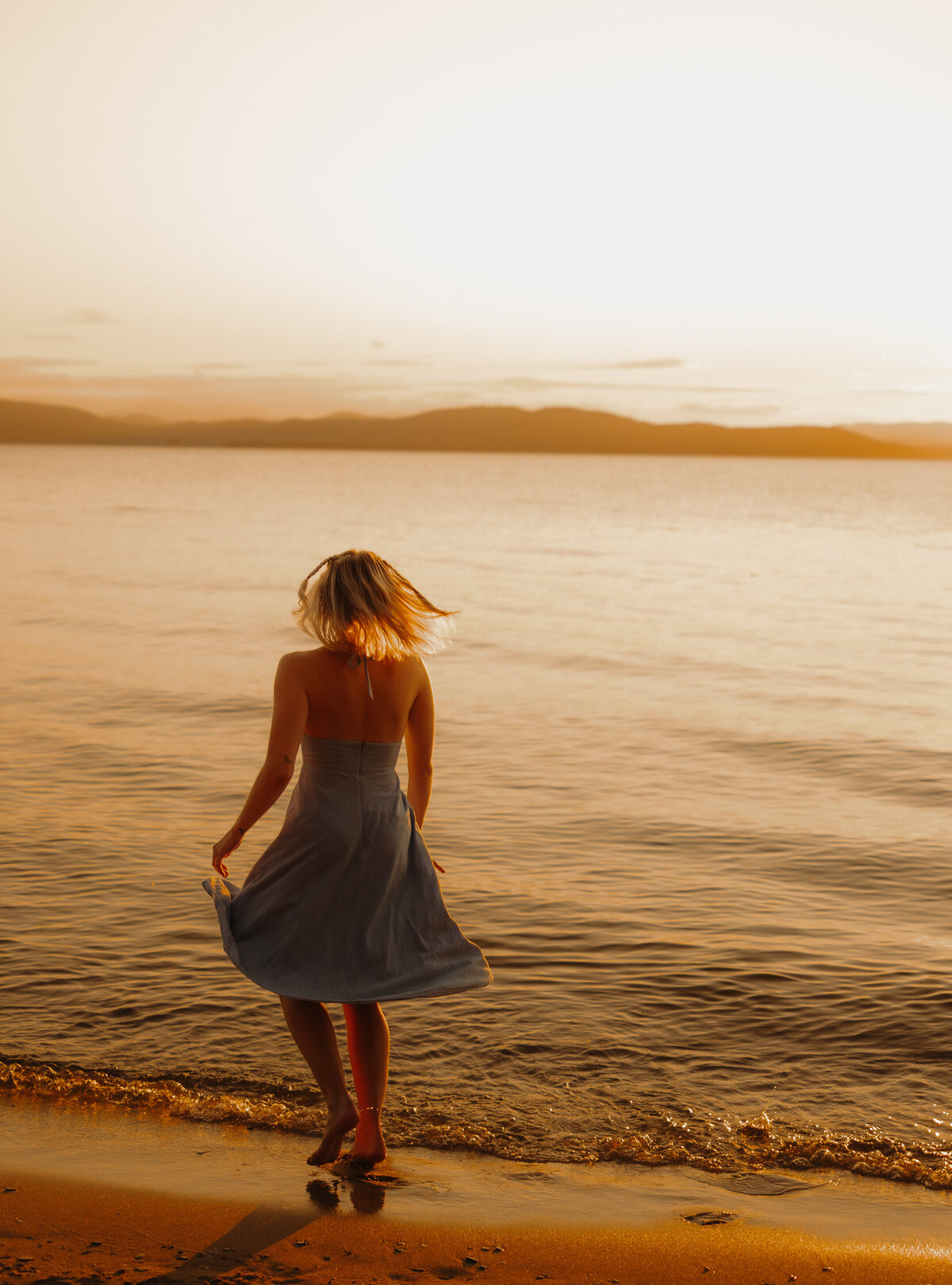 A woman with blonde hair in a blue dress stands at the edge of a sandy beach during sunset, captured by a New England portrait photographer. The sky and sea glow with warm golden tones, with distant hills gracing the horizon, creating a serene and timeless scene.