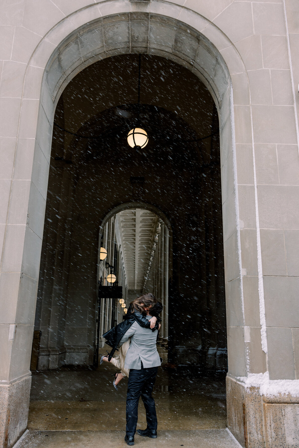 A couple framed under an arch outside Union Station in Chicago as snow falls, capturing a timeless and romantic winter engagement moment.