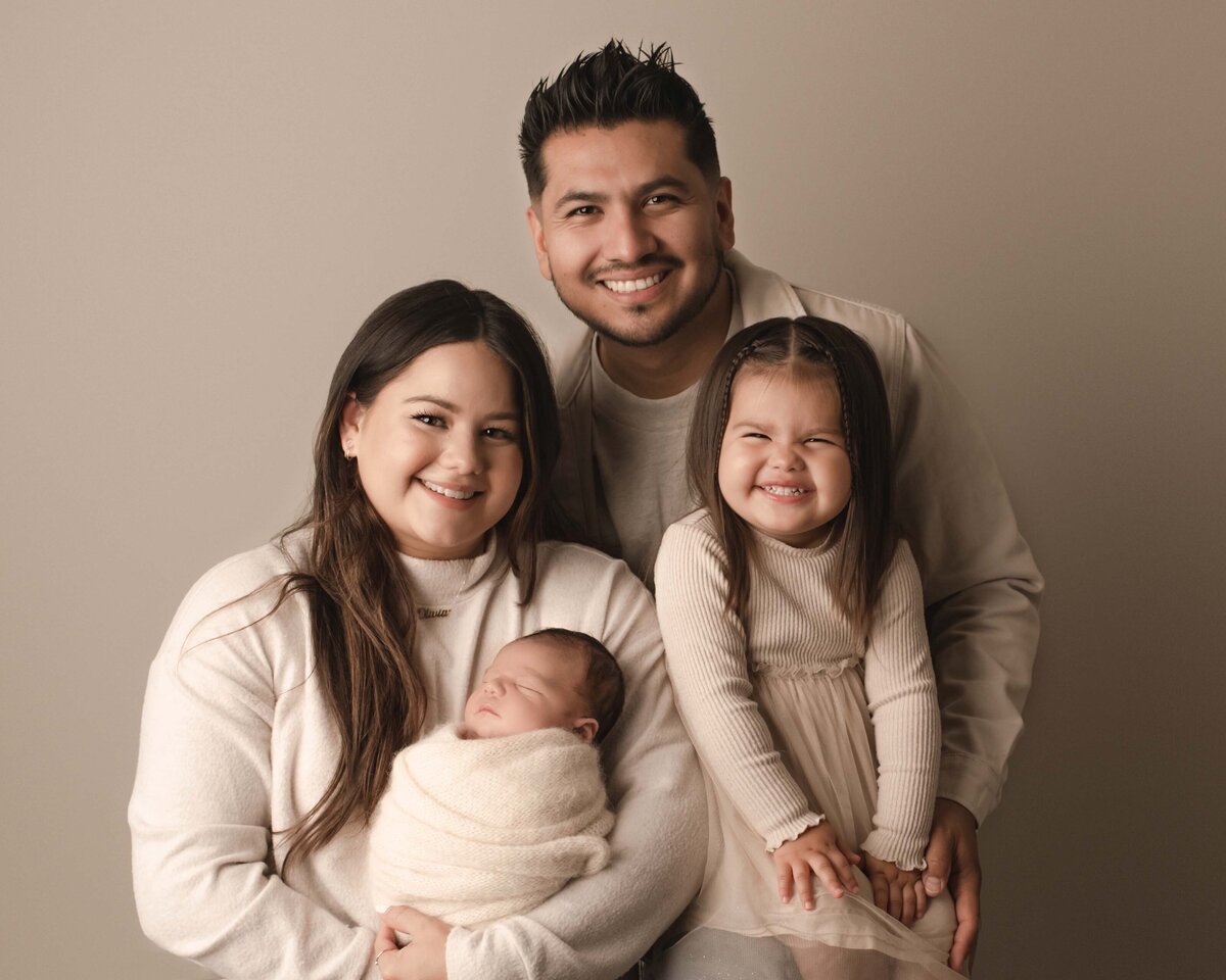 Mom, Dad and big sister pose with their newborn baby boy. Mom, dad, and big sister are smiling at the camera as mom is holding the sleeping baby boy. Captured by best Temecula newborn photographer Bonny Lynn Photography.