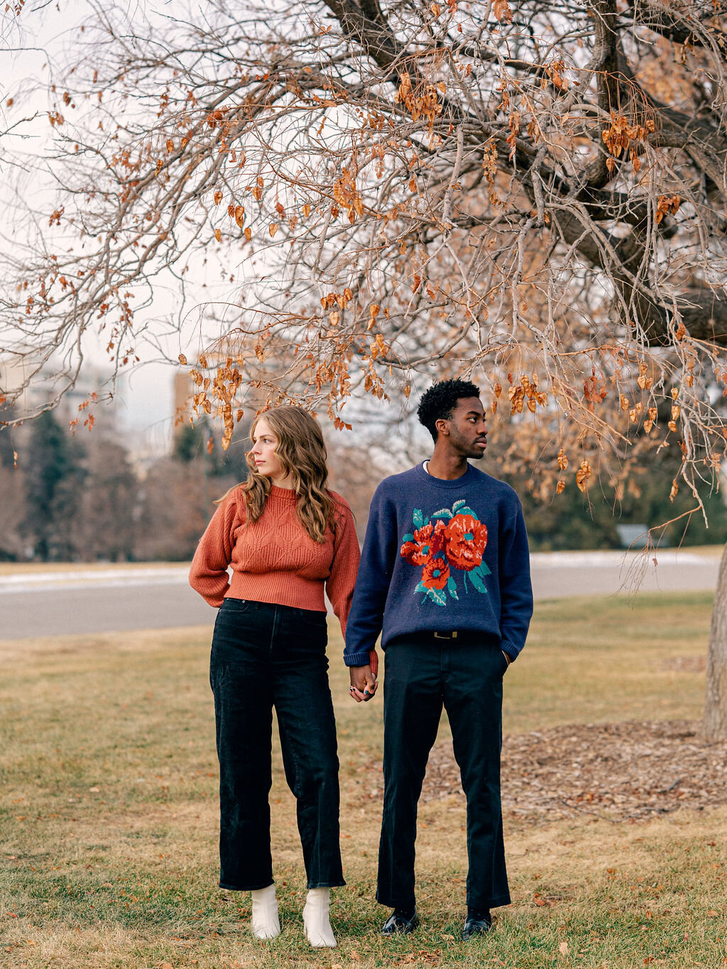 A couple stands under a tree at Cheesman Park. They are holding hands and looking in opposite directions.