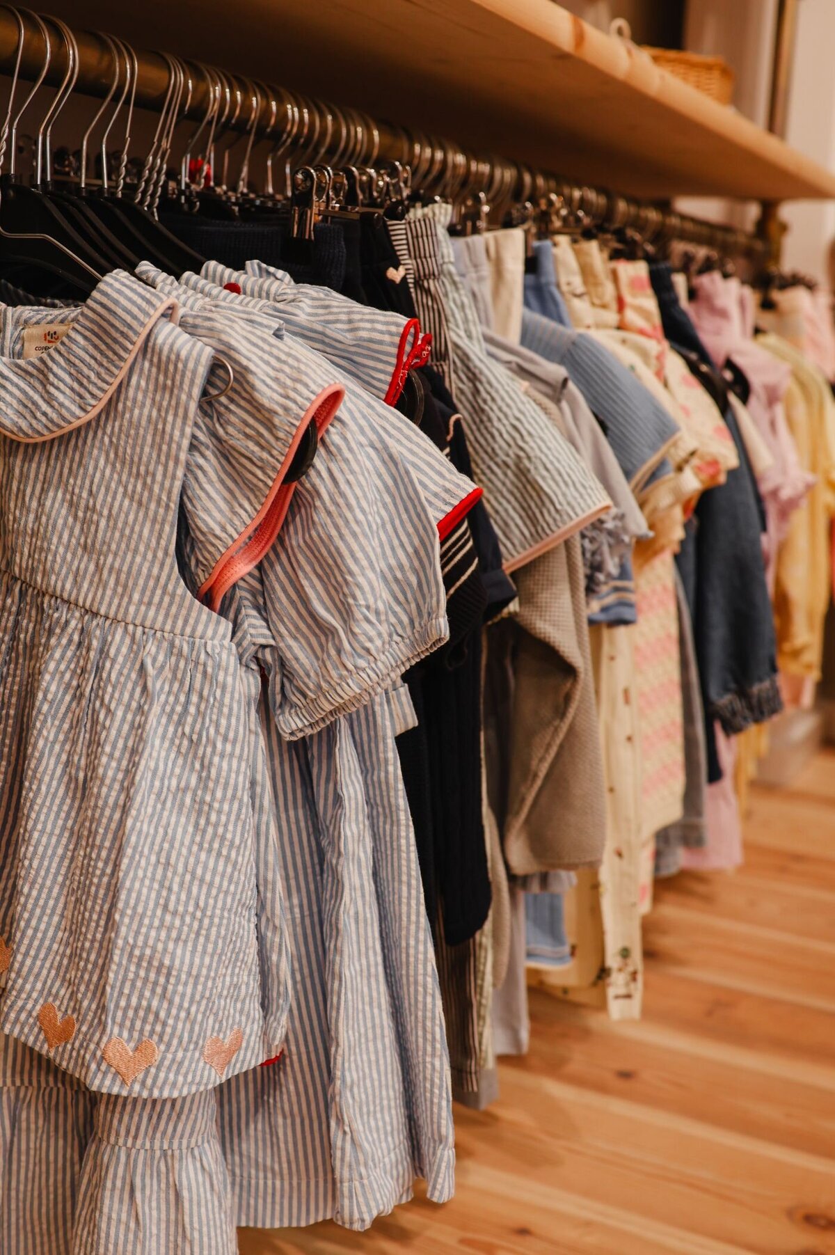 Close-up van een gestreept blauw-wit jurkje met korte mouwen en roze accenten, hangend aan een kledinghanger tussen andere kinderkleding in een winkel.