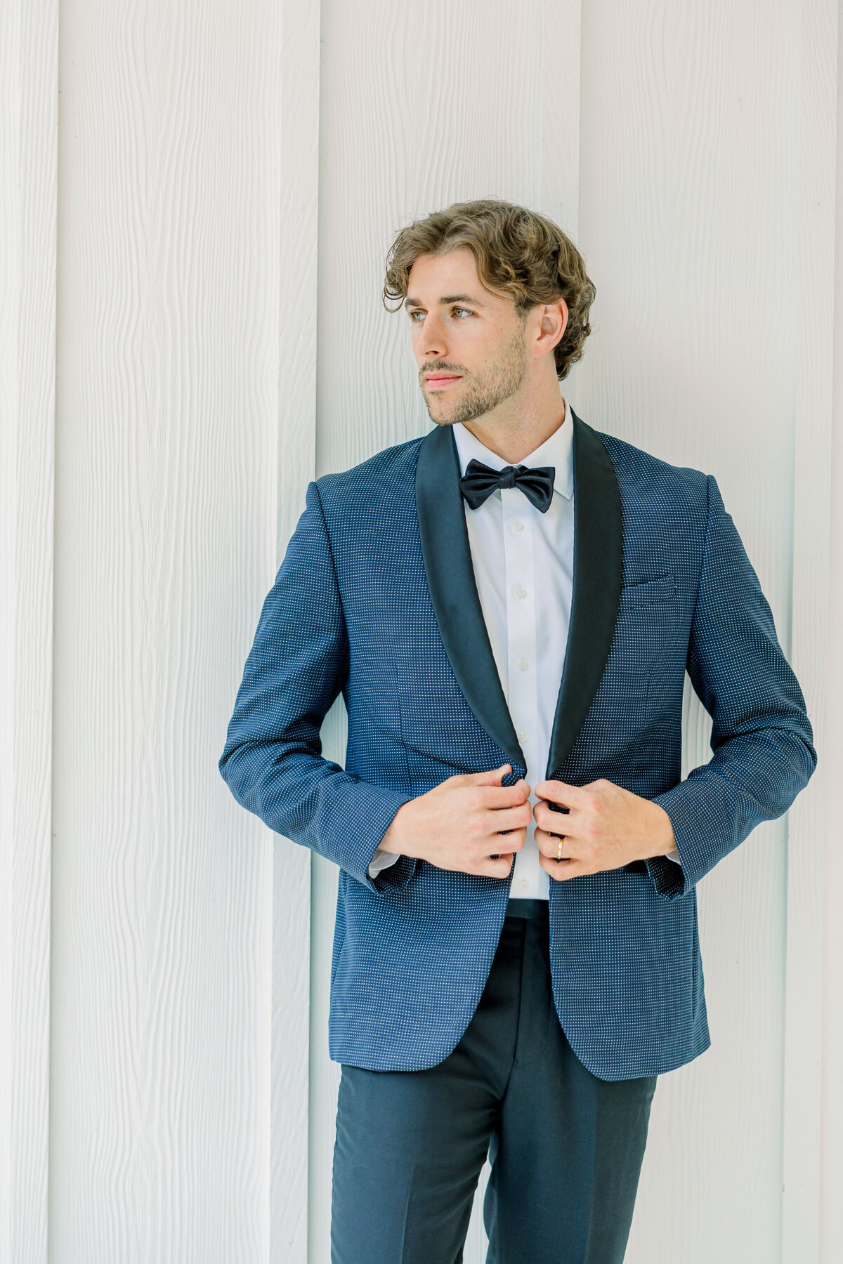 Ivy Rose Barn | A groom in a navy tuxedo buttons his jacket against the backdrop of a white wall at the Ivy Rose Barn, a white barn wedding venue in Virginia. Captured by Virginia Wedding Photographer, Caitlin Wilcox.