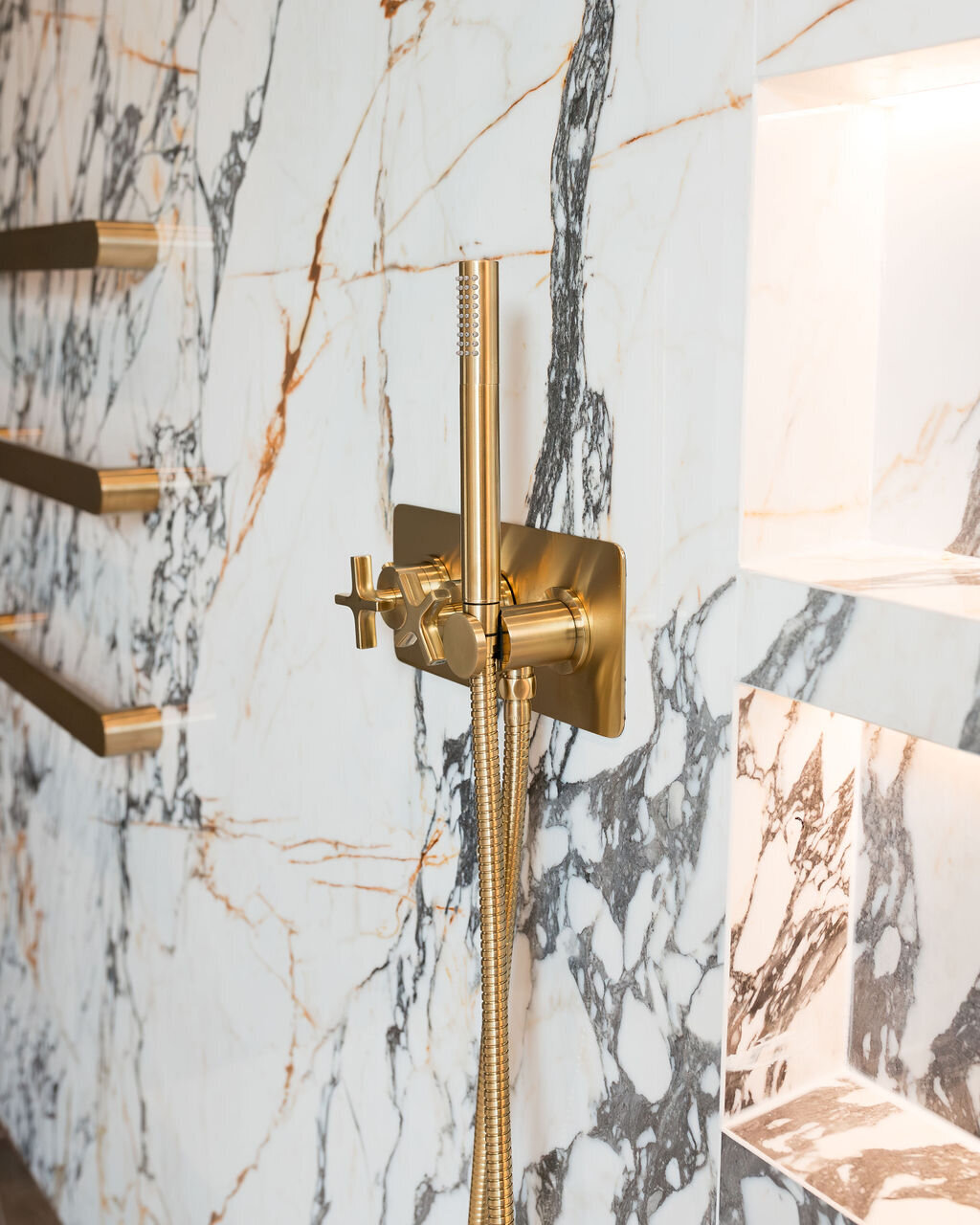 Modern gold towel bars and shower fixtures are mounted on a white marble wall with black veining.