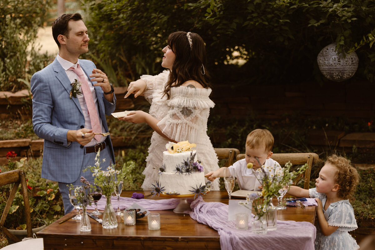 Bride and groom laugh while cutting their wedding cake during an intimate backyard celebration, with two young children seated at the table enjoying the moment.