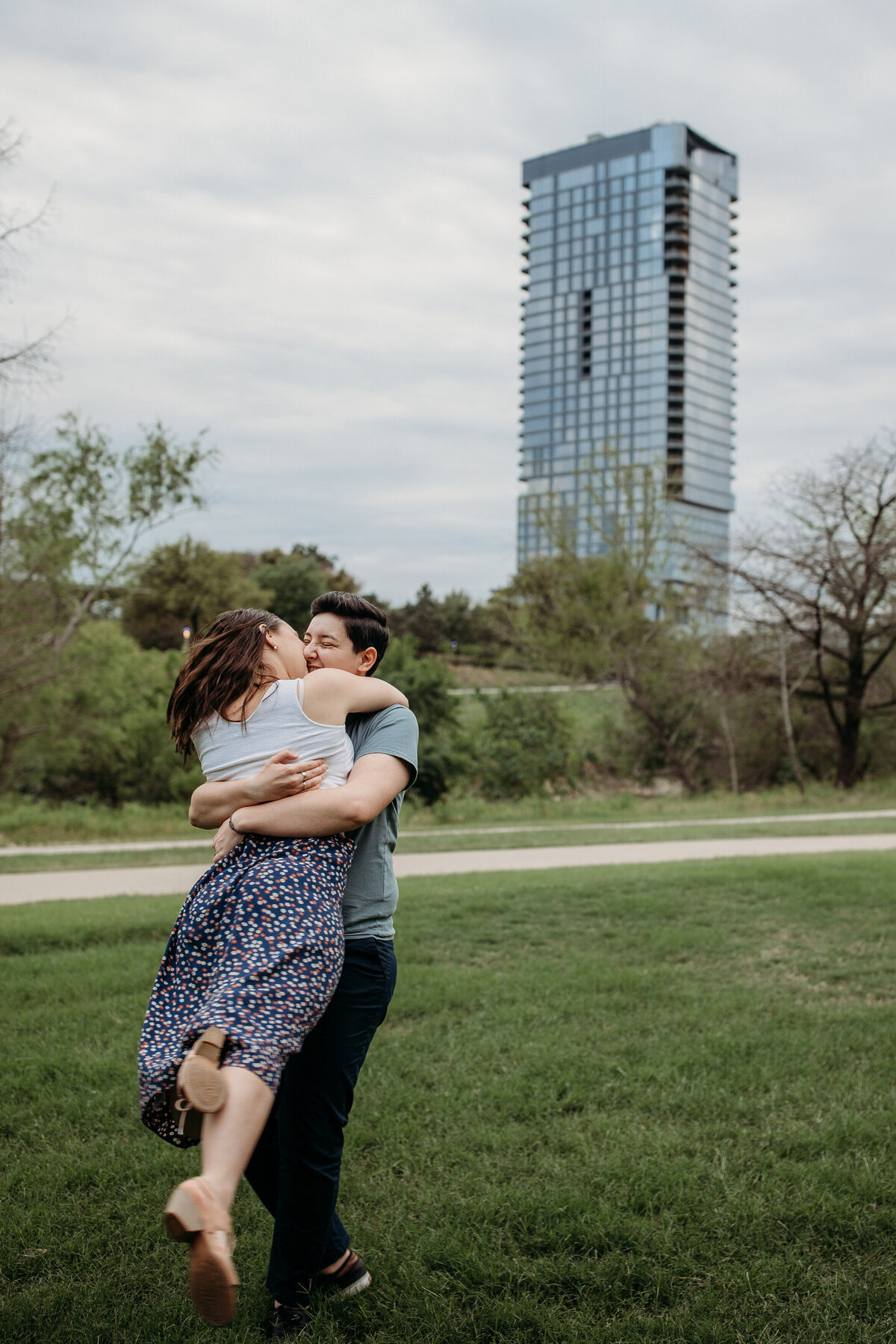 A couple in a joyful embrace, with one person lifting the other, set against a city park backdrop
